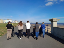 A photo taken from behind of a groups of mothers walking along a promenade. The sky is blur and the sea can be seen to the right hand side